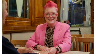 A woman with a bright pink blazer smiles as she sits at a table with another woman