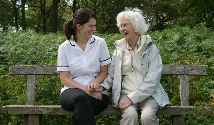 A nurse smiles and laughs with an older lady outside on a bench