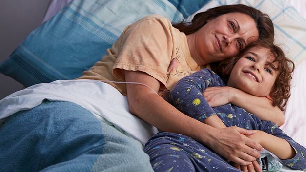 A mother and child are lying down in a hospice bed and smiling as they share a hug